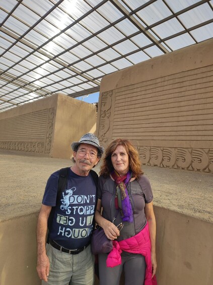 Two people standing in front of a large, textured wall with carved figures. 4