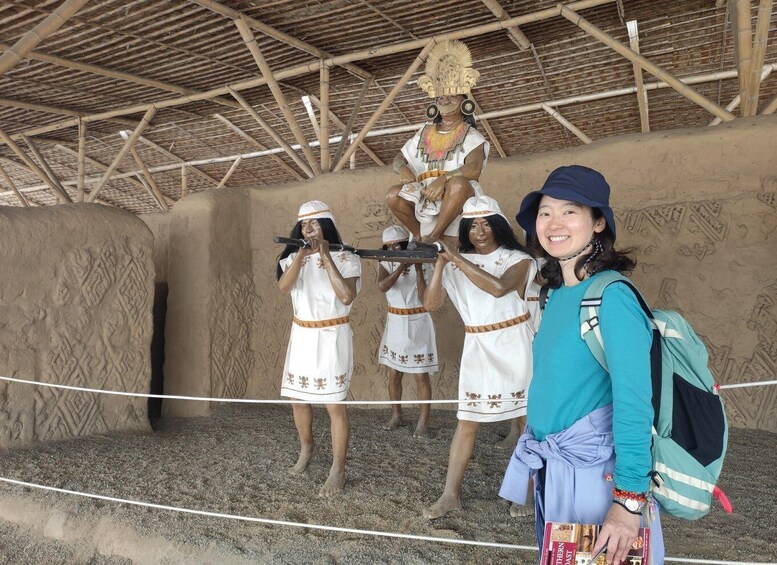 A person in a white dress with a headdress, holding a spear, and standing on a sandy surface with a rope barrier. 2