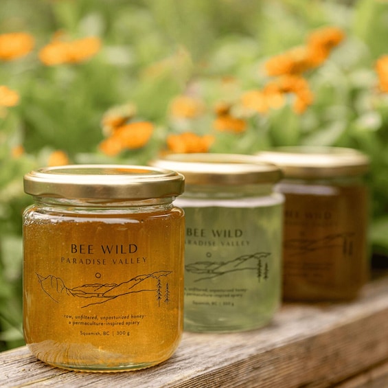 Three jars of Bee Wild honey on a wooden surface with a blurred background of greenery and orange flowers. 2