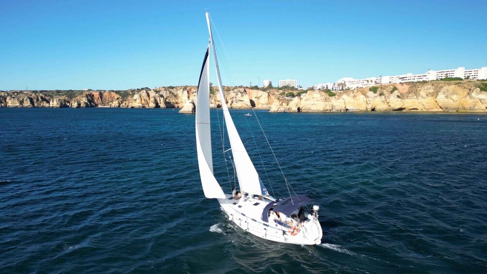 A sailboat on calm waters with a rocky coastline and buildings in the background. 3