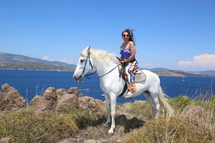A person riding a white horse on rocky terrain with a clear blue sea and distant mountains in the background. 3