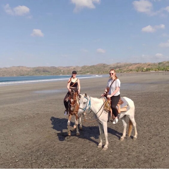Two people riding horses on a beach with a body of water and hills in the background. 4