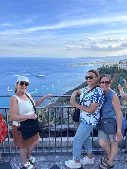 Three people taking a selfie with a scenic view of the sea and a distant cityscape. 2
