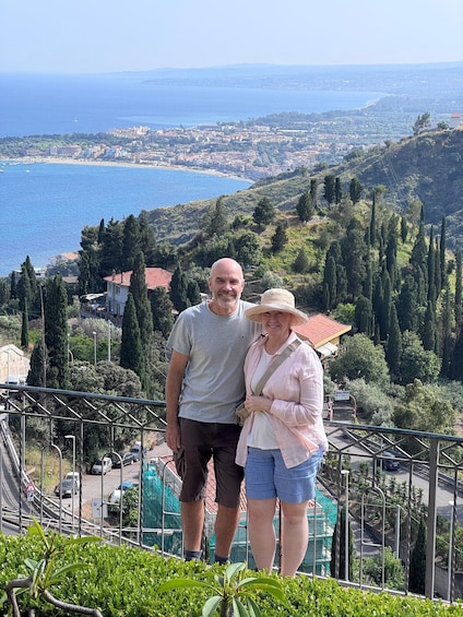 Two people standing on a viewing platform with a coastal landscape in the background. 1