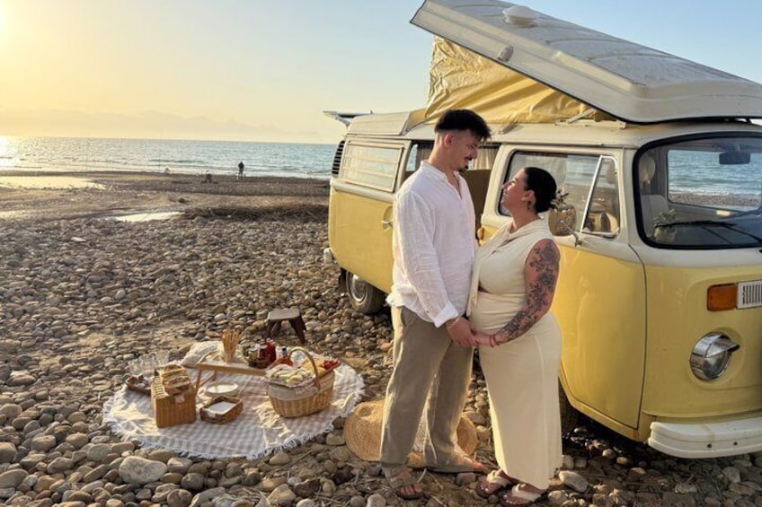 Un couple se tient près d’un vieux fourgonnet sur une plage rocheuse, à côté d’un coin pique-nique aménagé. 1