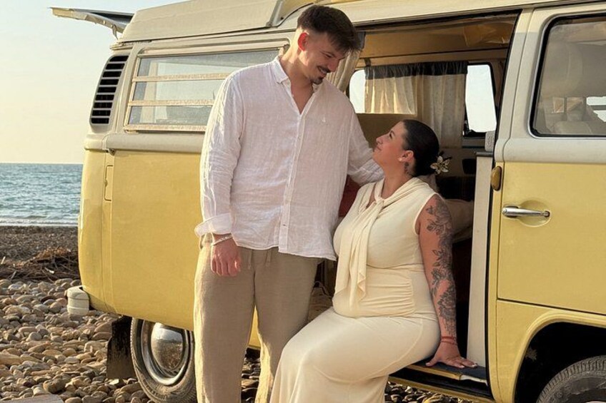 A couple standing next to a vintage van on a rocky beach with the ocean in the background. 2
