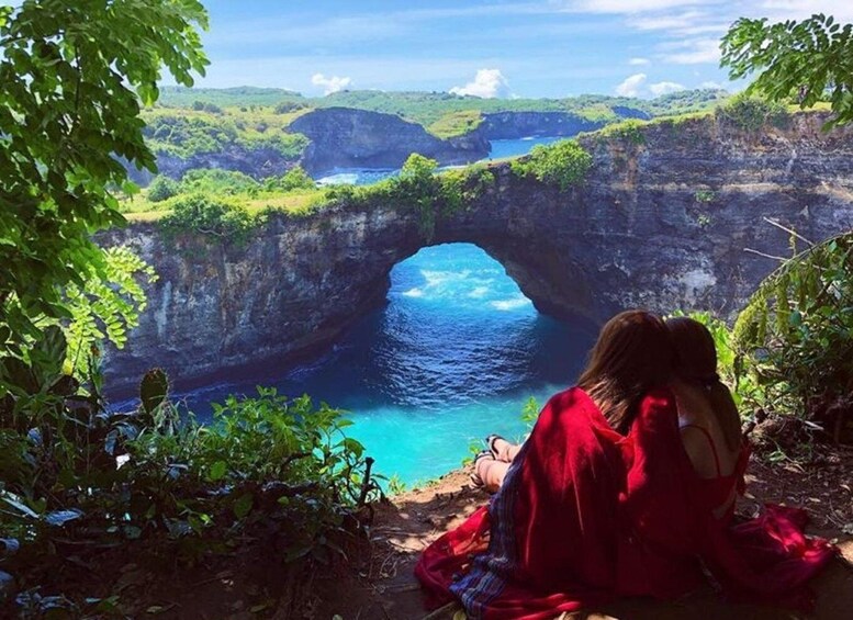 A woman in red sitting by a natural archway over a blue body of water. 1