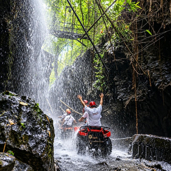 A person on a red ATV navigating through a waterfall in a forested area. 1