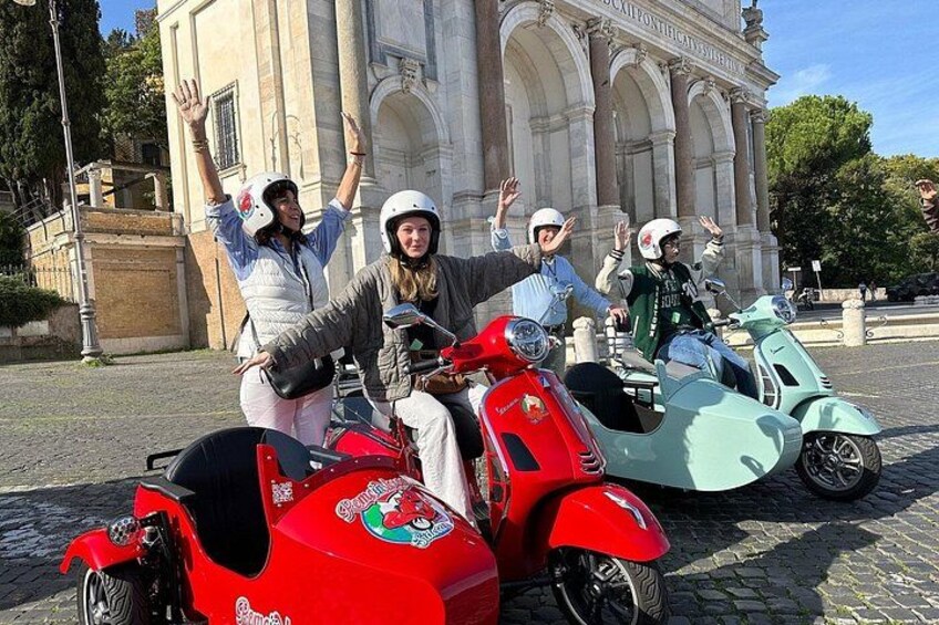 Four people on vintage scooters in front of a historic building. 1