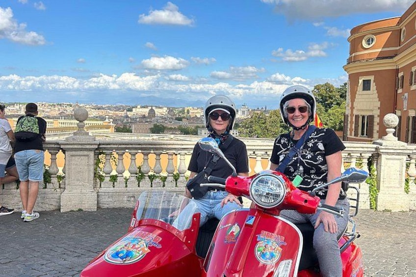 Two people on a red scooter with a cityscape in the background. 2