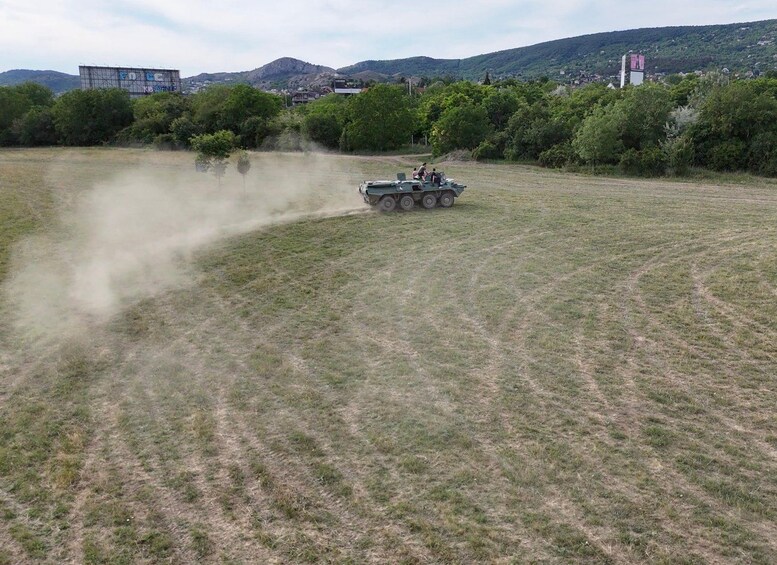 A military tank moving across a field with dust being kicked up, surrounded by greenery and hills in the background. 2