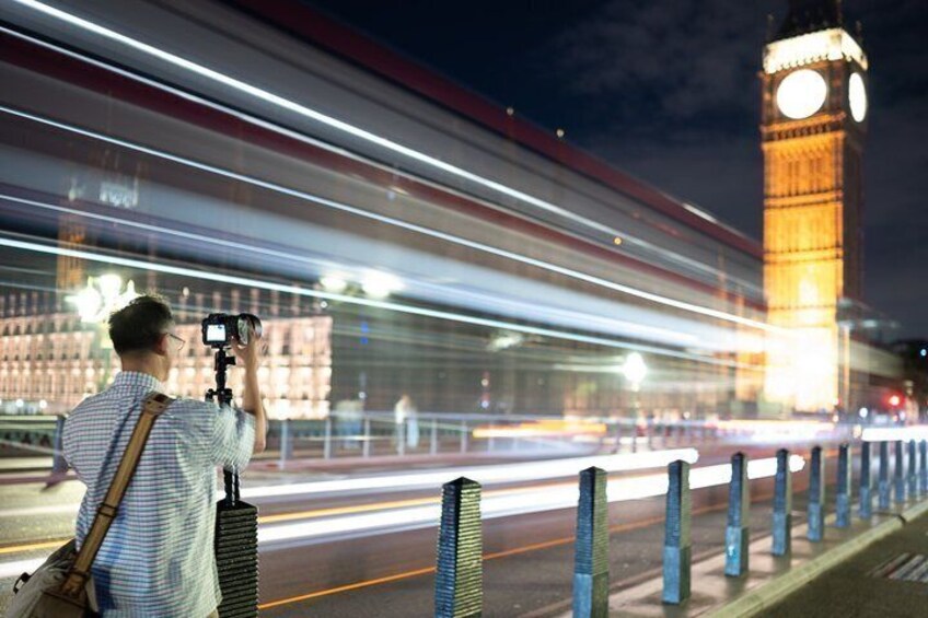 Une personne prenant une photo avec un appareil photo, une grande tour de l’horloge et des lumières floues provenant d’un véhicule en mouvement. 1