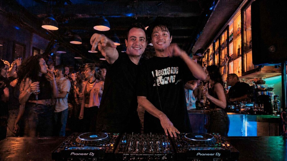 Two people standing behind Pioneer DJ equipment in a dimly lit bar with people dancing in the background. 4
