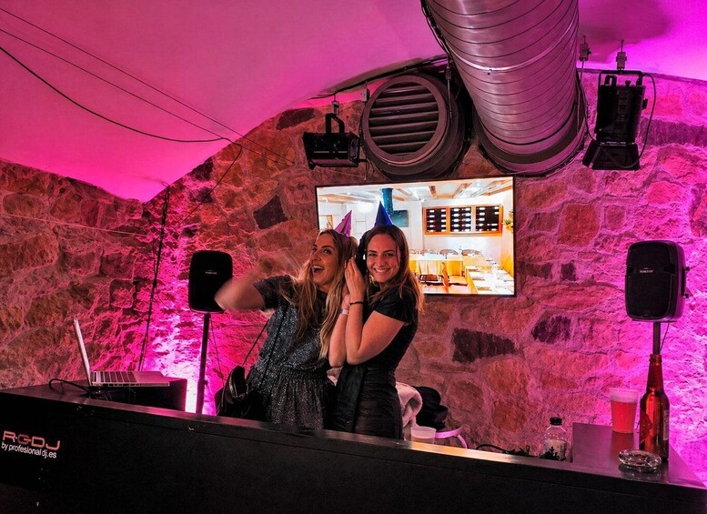 Two people at a DJ booth with a stone wall background and a television screen. 2
