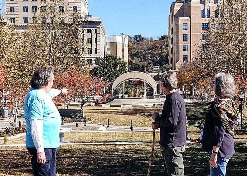 Three people standing in a park with autumn foliage, a stone bridge, and buildings in the background. 2