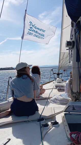 En sejlbåd med et hvidt flag, der viser 'Velanore Siracusa C320 0406155', og to personer sidder på dækket. 2