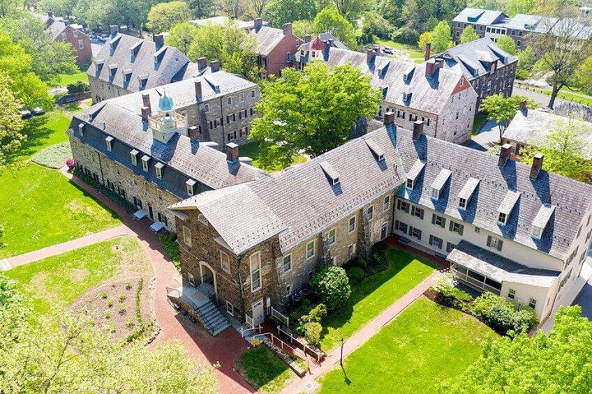 A large, historic building with a steeply pitched roof, surrounded by lush green lawns and smaller buildings. 3