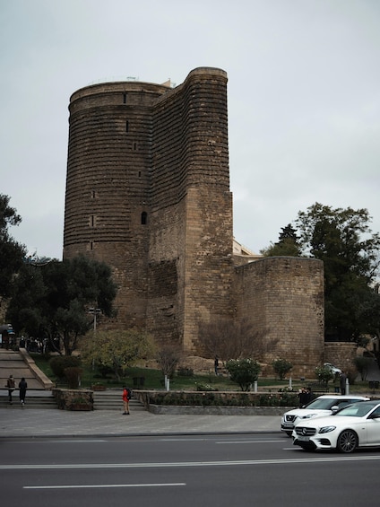 A historic stone tower with a curved top, surrounded by trees and a paved area with people and cars. 2