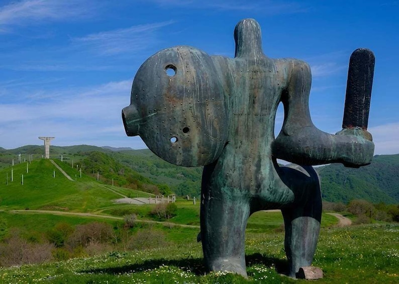 A large bronze sculpture of a humanoid figure with a cylindrical shape on its right side, set against a backdrop of green hills and wind turbines. 5