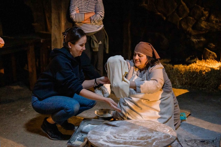 Two people kneeling on the ground, one teaching the other how to cook. 1