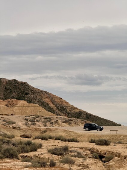 A black SUV driving on a dirt road in a desert landscape with sparse vegetation and a mountain in the background. 4