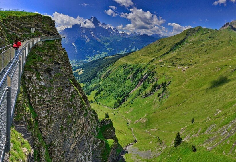 A suspension bridge over a steep cliff, a valley with green hills, and distant mountains under a blue sky. 2