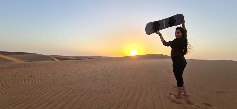 A person holding a snowboard in a desert landscape with sand dunes and a sunset in the background. 1