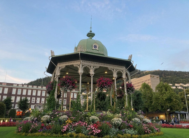 A gazebo with a green dome, surrounded by a flower garden and buildings in the background. 1