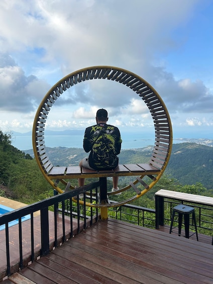 A person sitting on a bench with a circular metal frame, overlooking a mountainous landscape. 2