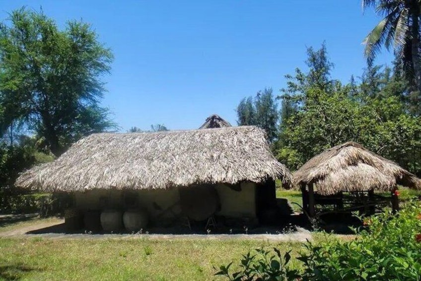 A traditional thatched-roof house with a small adjacent hut, surrounded by lush greenery and palm trees. 4