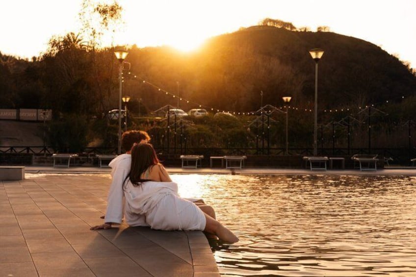 Two people sitting by a water body during sunset, with a hilly landscape and street lamps in the background. 1
