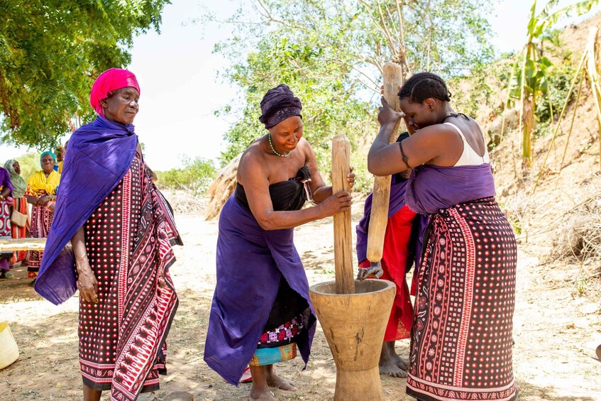 Three individuals engaged in a traditional activity, possibly grinding or pounding, with a large wooden mortar and pestle. 3