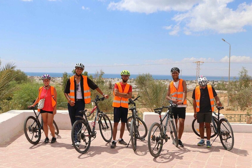 A group of cyclists in safety vests standing with bicycles on a paved area with a coastal view. 2