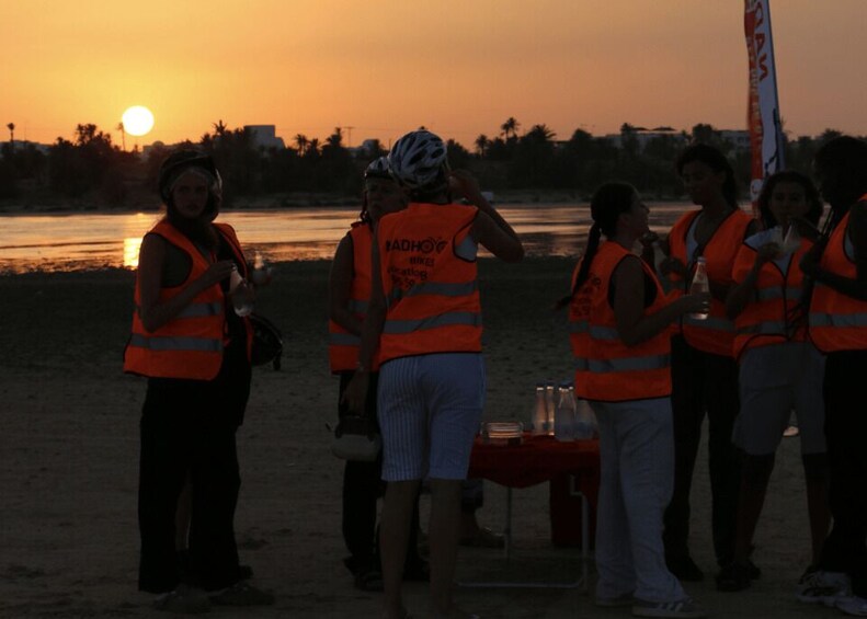 A group of people in orange vests standing on a beach at sunset. 1