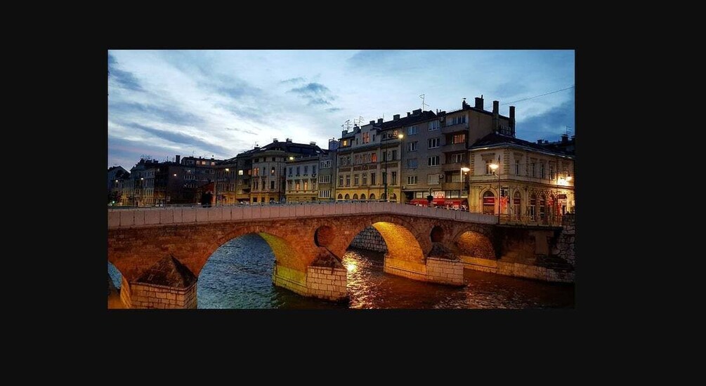 A stone bridge with arches over a body of water, surrounded by buildings with illuminated windows. 1