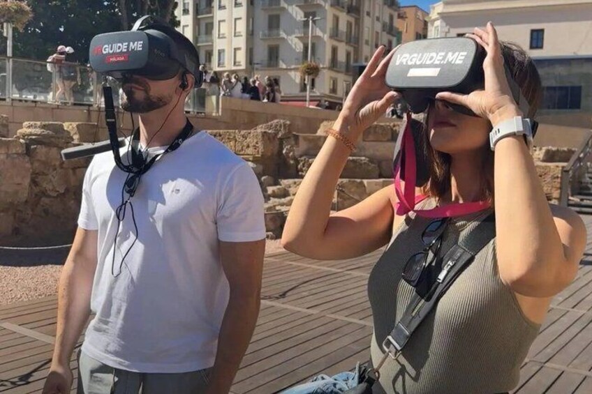 Due persone indossano visori VR con la scritta 'VRGUIDE ME', in piedi su una passerella di legno con edifici sullo sfondo. 4