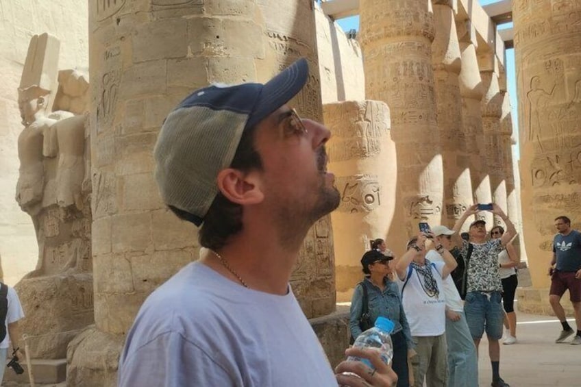 A person wearing a cap and holding a water bottle, standing in front of ancient Egyptian columns with hieroglyphs. 5