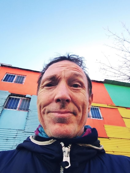 A person in a dark jacket with a zipper, standing in front of a colorful building with red, green, and blue sections. 3