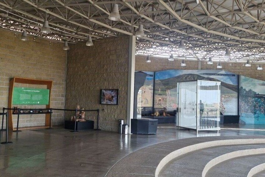 An indoor exhibition space with a brick wall, a green informational board, a sculpture, and a mural. 2