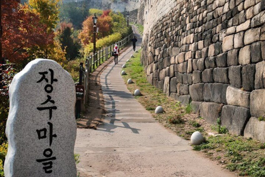 En stensti med en stenvæg på den ene side, en marksten med koreansk tekst, og en person der går i det fjerne. 2