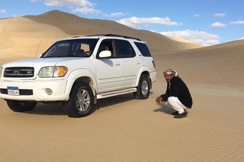 A white SUV parked on a sandy dune with a person crouching beside it. 1