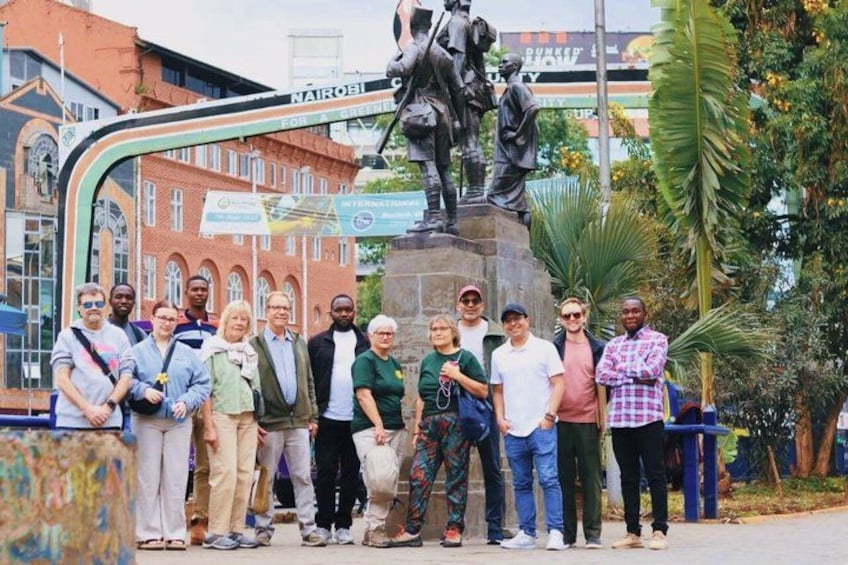 Un groupe de personnes posant devant une statue, avec en arrière-plan des bâtiments et une pancarte sur laquelle on peut lire « NAIROBI ». 1