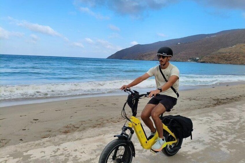Une personne fait du vélo sur une plage de sable, à bord d’un vélo électrique jaune, avec l’océan et des collines en arrière-plan. 4