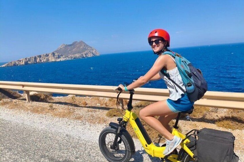 Une personne à vélo, de couleur jaune, porte un casque rouge et un sac à dos. Elle roule sur une route côtière, avec une île rocheuse en arrière-plan. 2