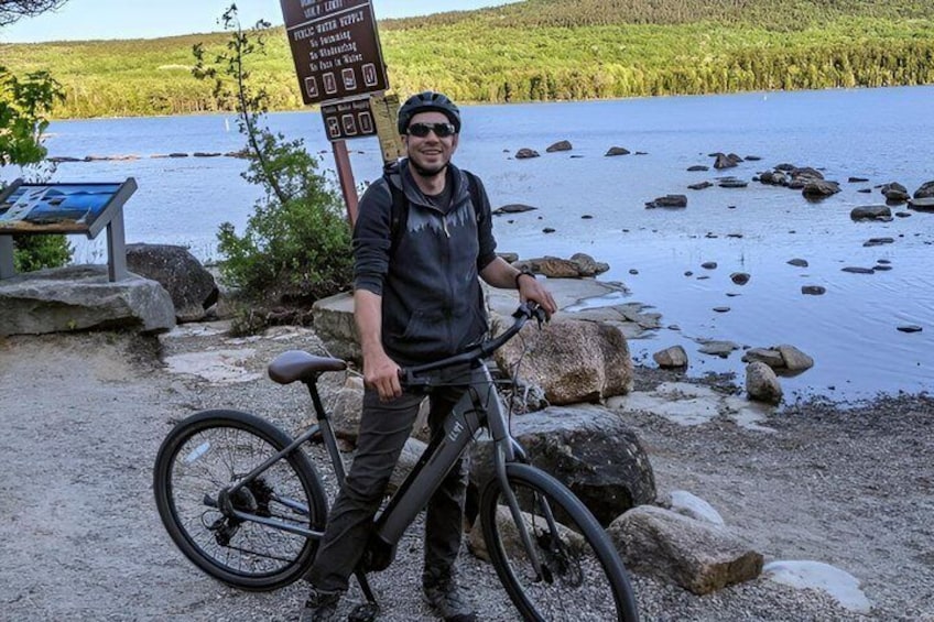 A person standing next to a bicycle by a lake with a signpost in the background. 4