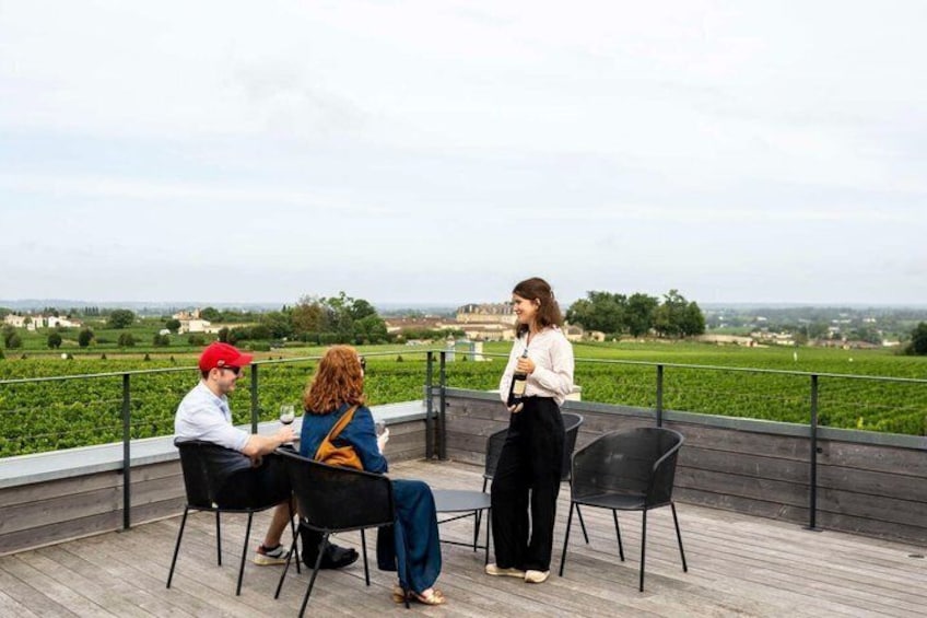Three people seated at a table on a wooden deck overlooking a vineyard. 3