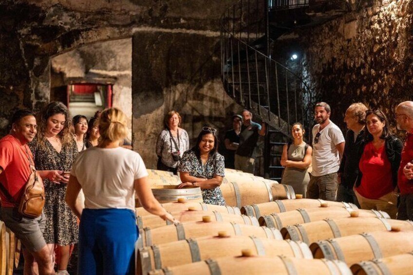 A group of people standing around large wooden barrels in a dimly lit room with stone walls and a spiral staircase in the background. 1
