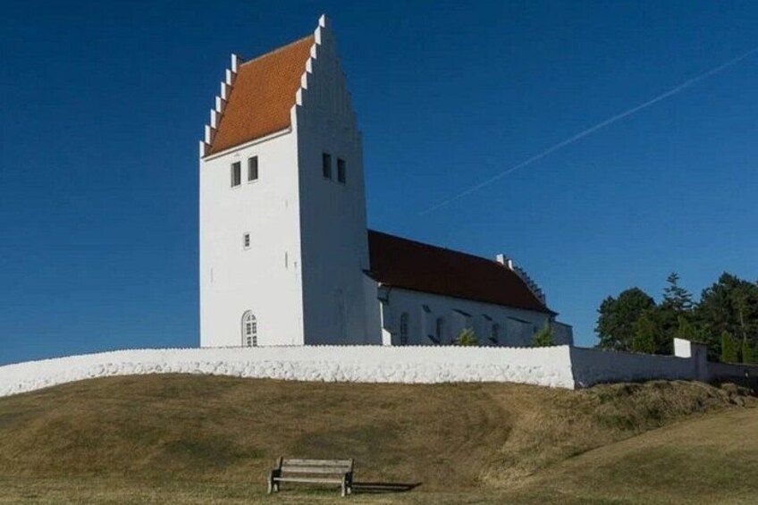 Una iglesia blanca con tejado rojo y un campanario alto, rodeada por un muro blanco y una colina cubierta de hierba. 5
