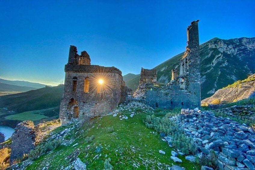 En historisk slottsruin av sten, med en nedgående sol i bakgrunden, omgiven av gröna kullar och berg. 1