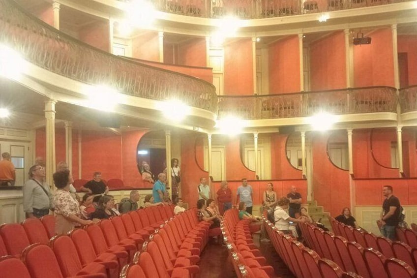 Un teatro con posti a sedere rossi e balconi, persone sedute e in piedi, e un'area palco. 3